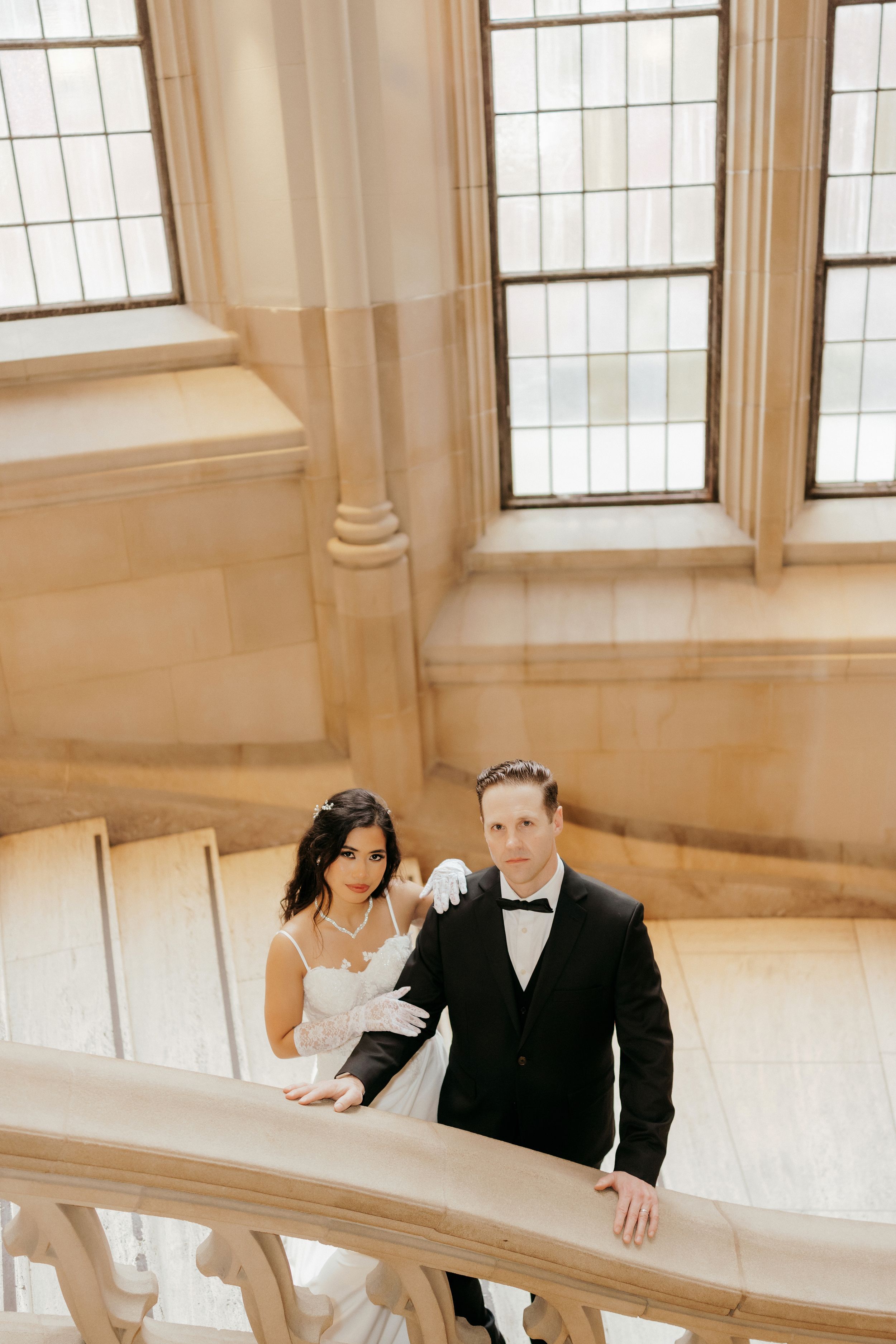 A romantic wedding couple poses along a curved marble staircase in an elegant venue with tall windows.