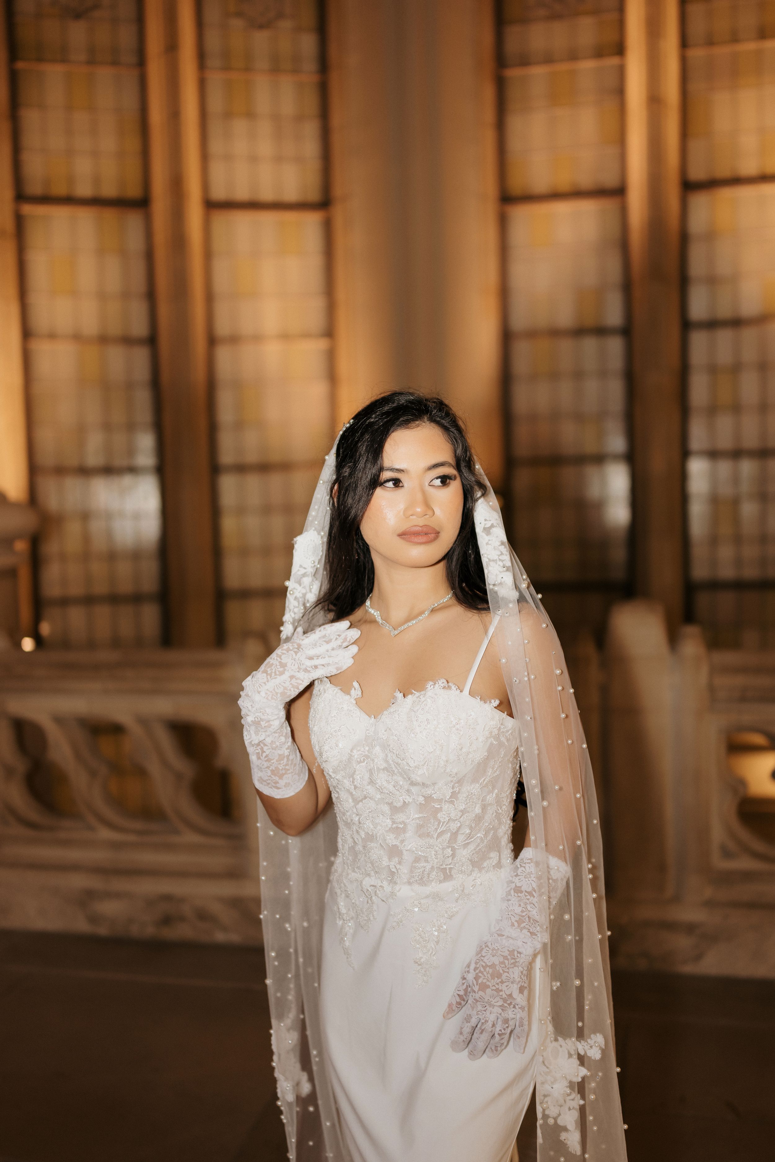A bride in a white lace wedding gown and veil stands elegantly in front of ornate windows in a historic venue.