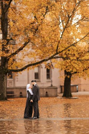 A romantic couple embraces beneath golden autumn trees on a leaf-covered ground near a stone building.