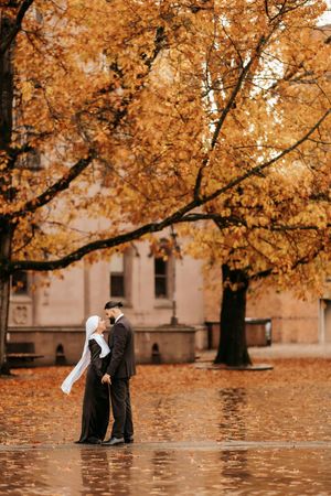 A couple shares an intimate moment under golden autumn trees with fallen leaves scattered on the ground.