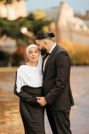 Romantic couple in black and white attire embrace during outdoor photoshoot with autumn scenery.