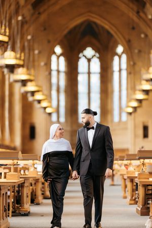 A couple walks hand in hand down the aisle of a grand cathedral with gothic arches and wooden pews.