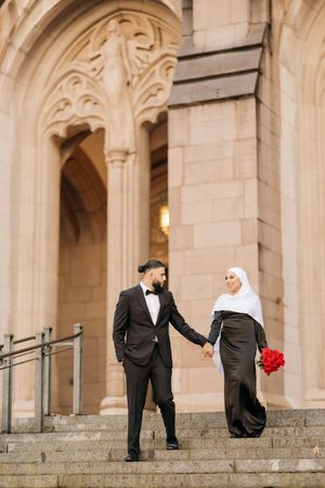 Wedding party members in black suits and red bouquets pose playfully on stone steps of an ornate architectural building.