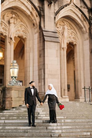 A couple in formal attire holding hands on stone steps of a grand Gothic-style building with ornate arches.