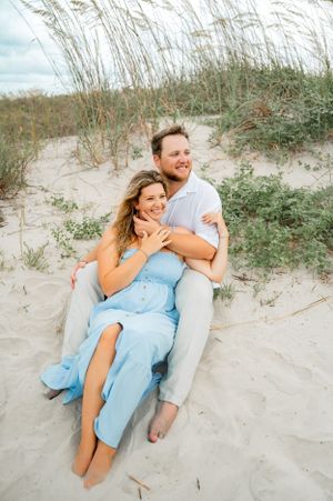 A couple shares a playful moment while sitting in beach sand dunes with sea grass.