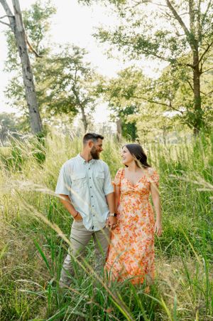 Couple walking together through tall grass and trees in golden afternoon light.