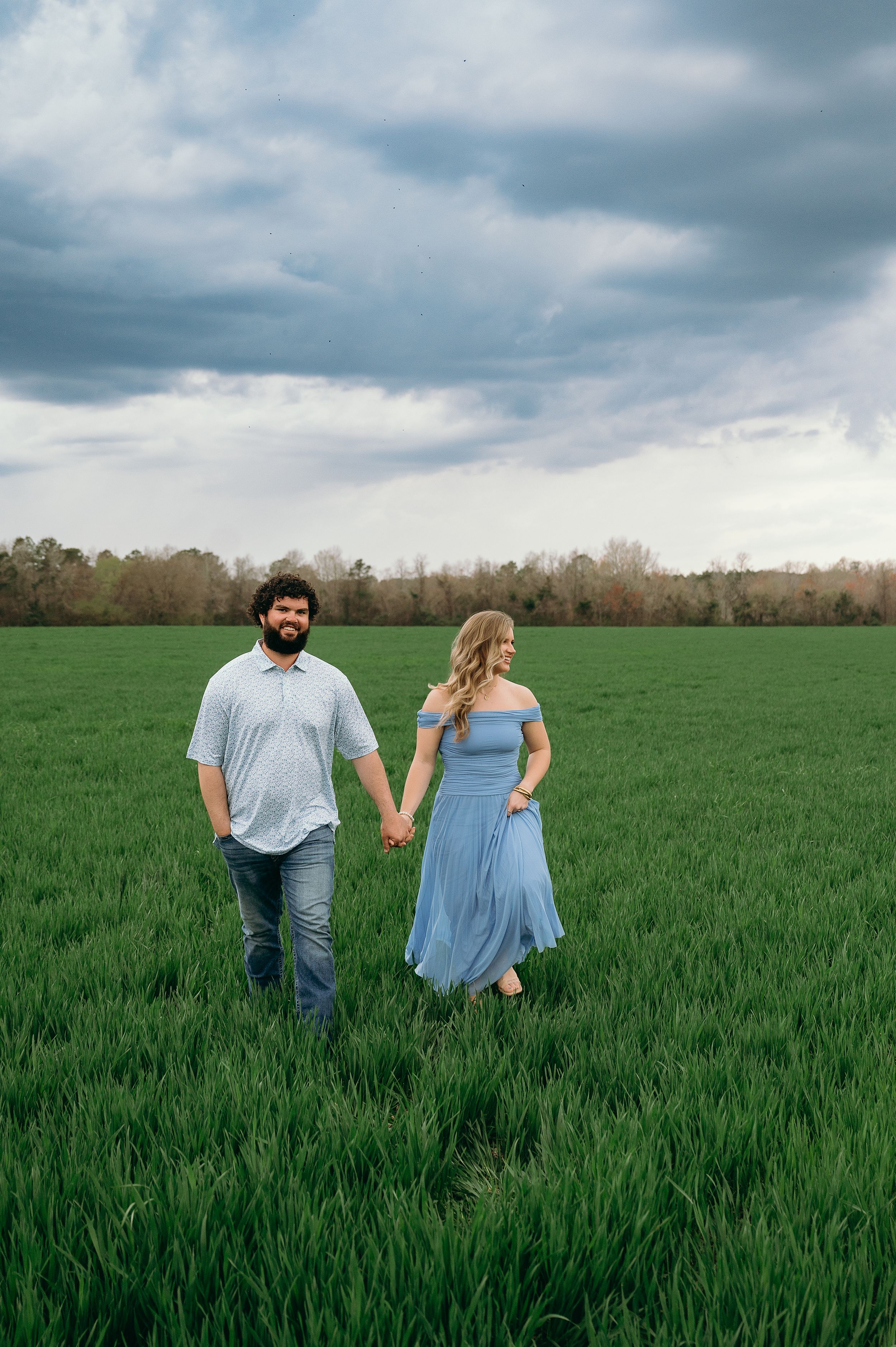 A couple holds hands walking through a lush green field under dramatic cloudy skies, wearing coordinating blue outfits.