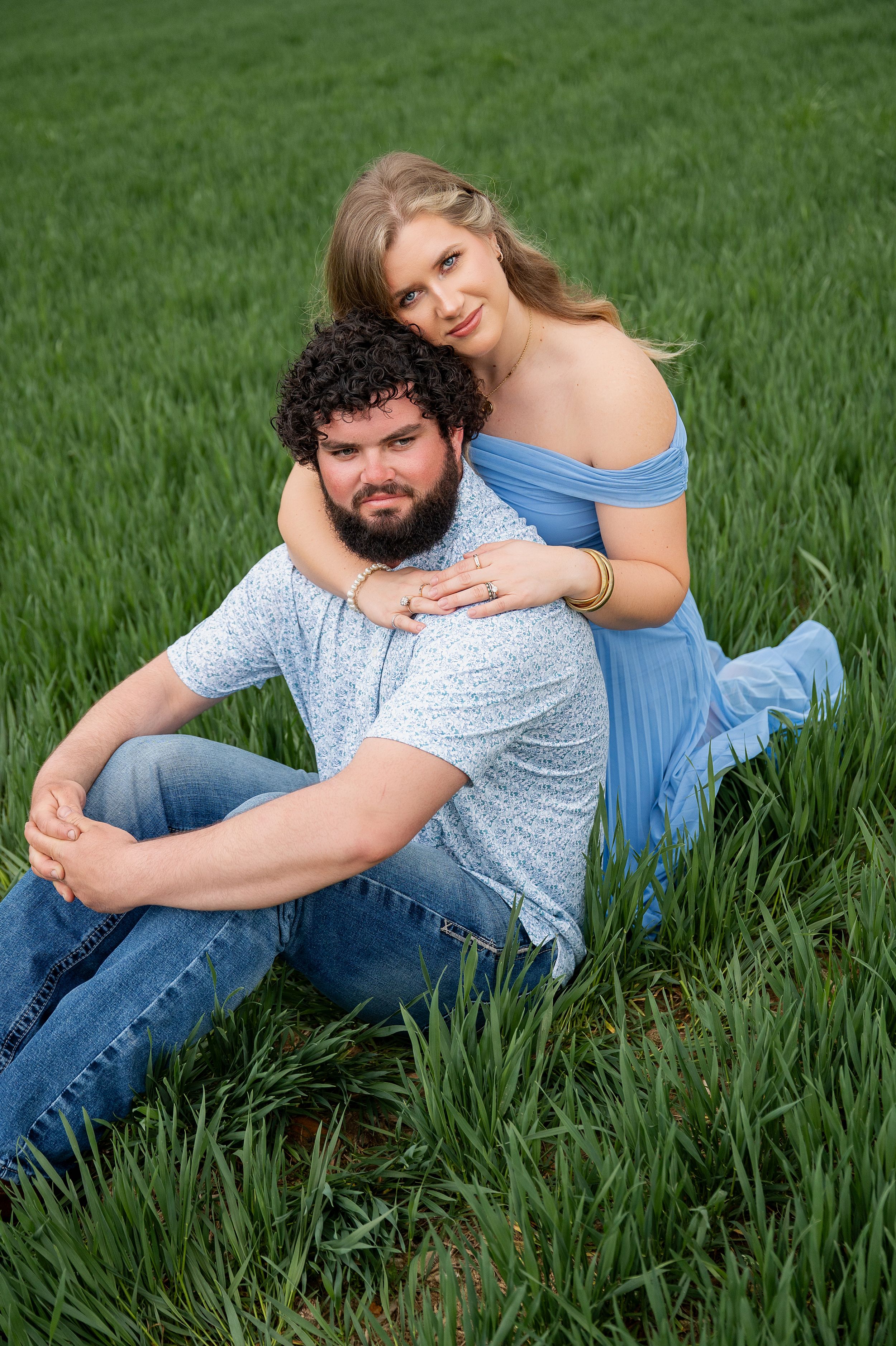 A couple poses in a lush green field, woman in blue dress embracing man with curly hair and beard sitting in grass.