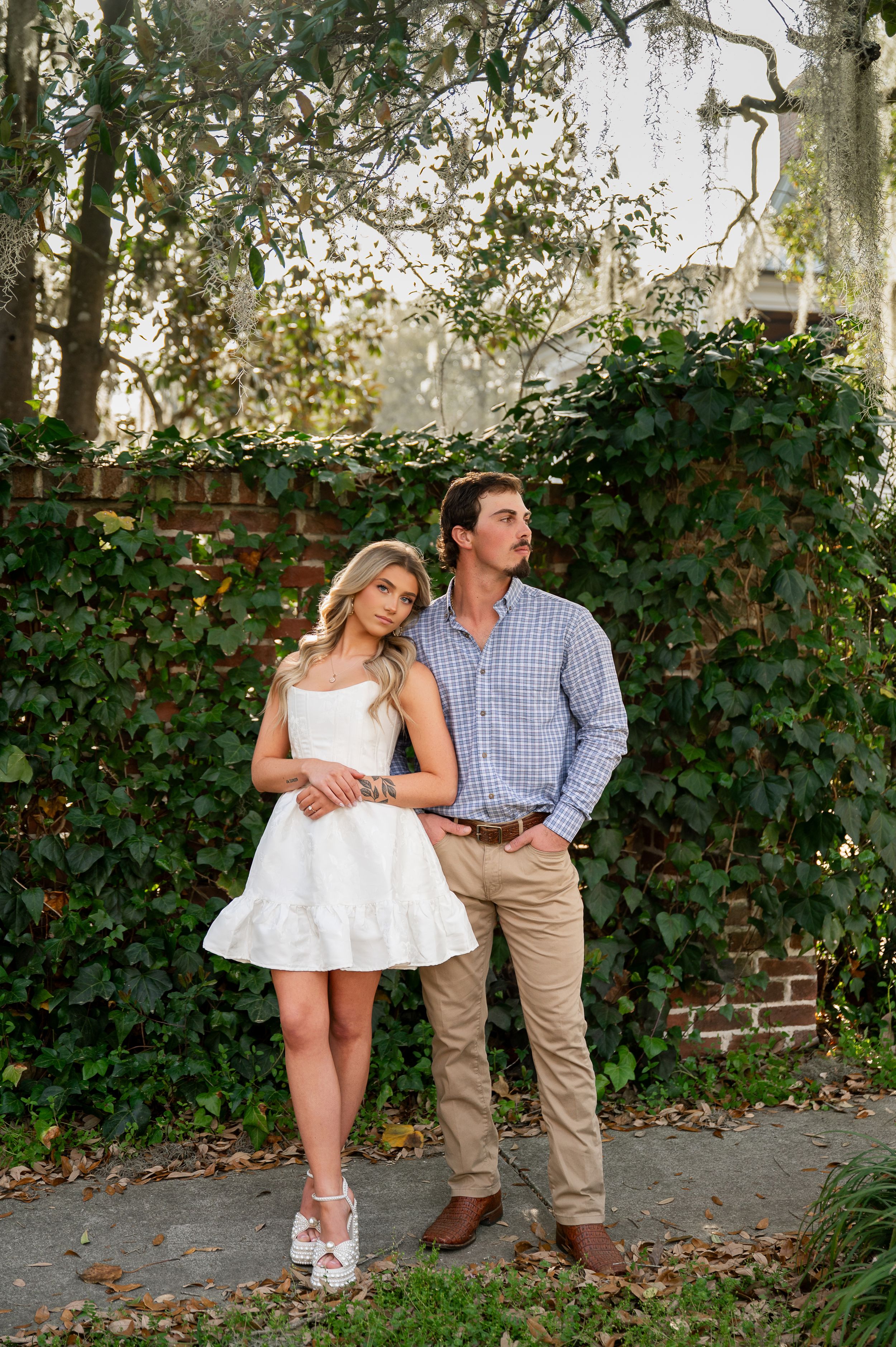 Couple posing outdoors against ivy-covered brick wall, woman in white mini dress, man in blue shirt and khakis.