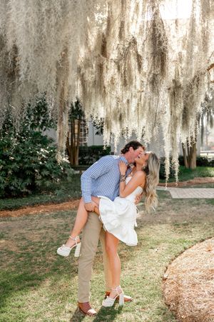 A man lifts and kisses a woman in a white dress under Spanish moss-draped trees during an outdoor engagement session.