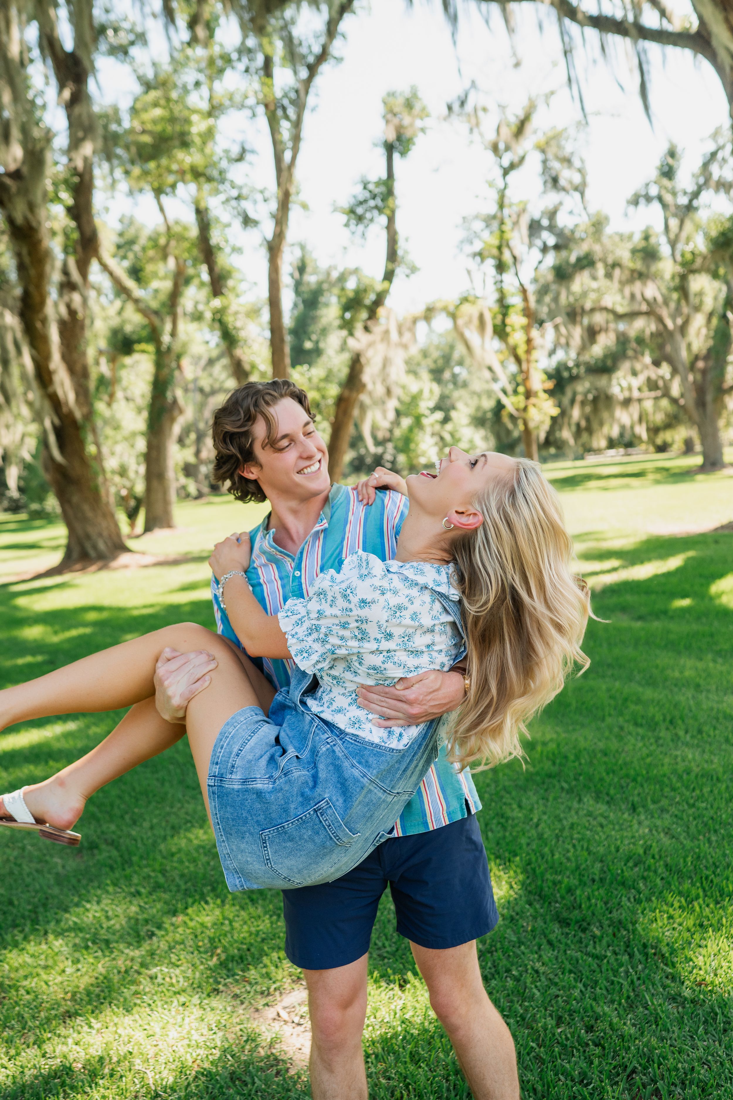 A smiling man carries a laughing blonde woman in a sunny green park with moss-draped trees.