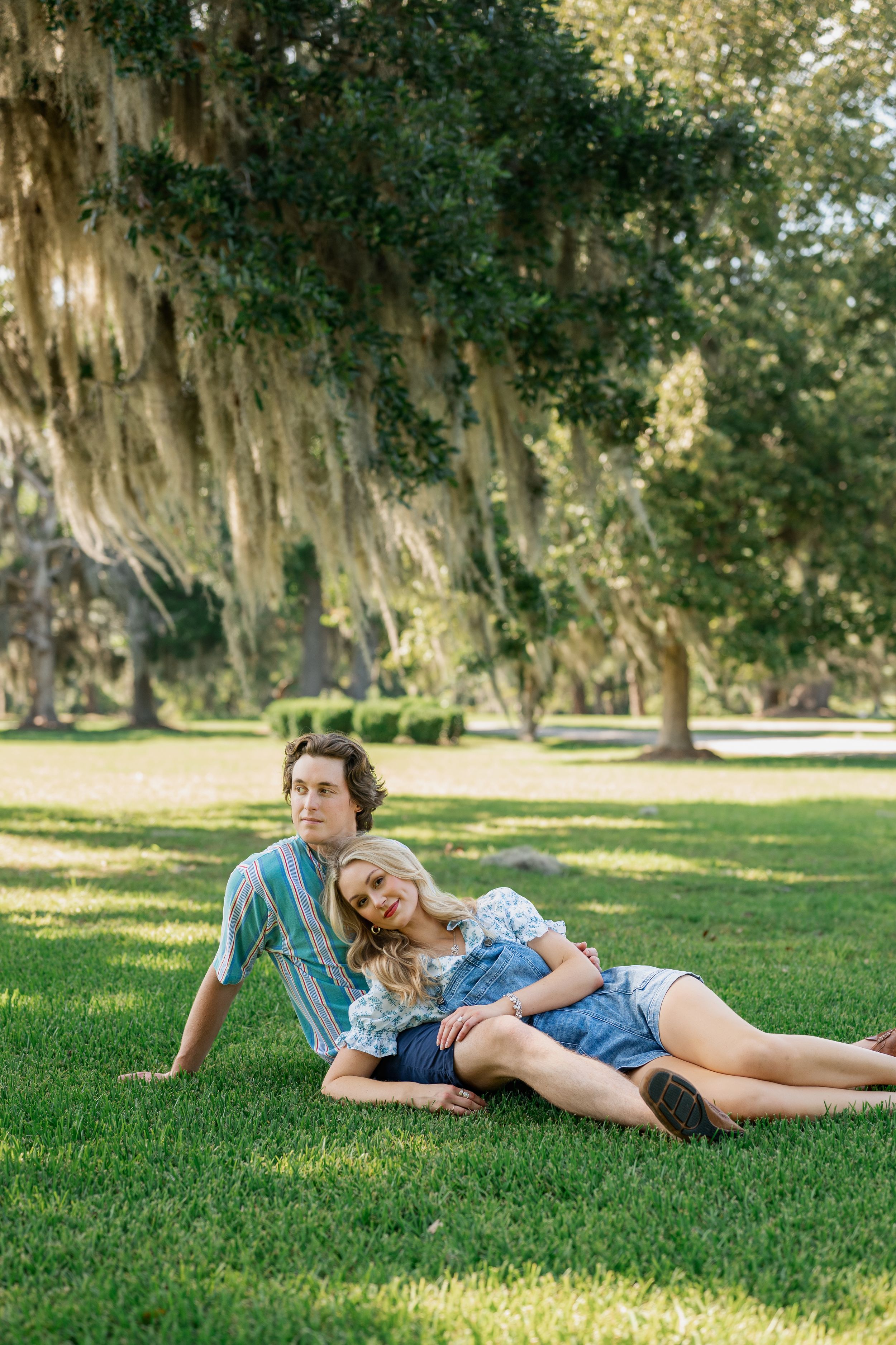 Couple relaxing on green grass under Spanish moss-draped oak trees in a sunny park setting.