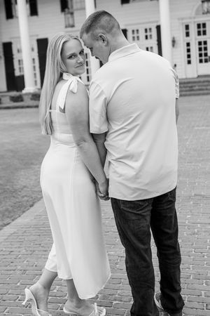 Black and white photo of a couple holding hands, woman in white dress glancing back, man in polo and jeans, elegant building behind.