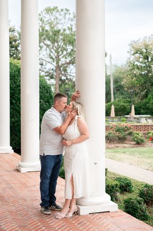 Couple sharing a romantic kiss between white columns in a garden setting, woman in white dress and man in casual attire.