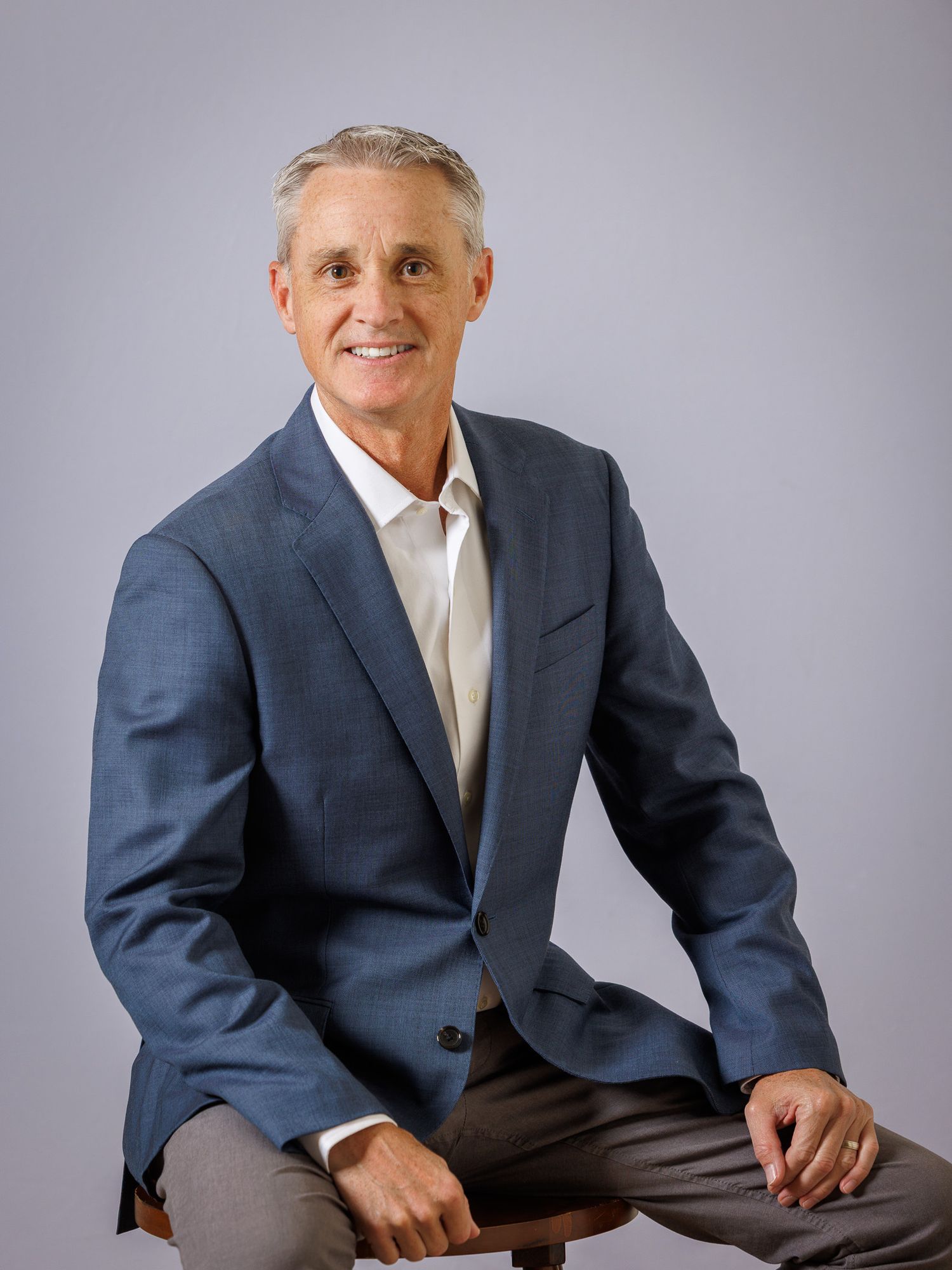 Professional wearing a navy suit and light tie sits in multiple poses against a gray studio background.