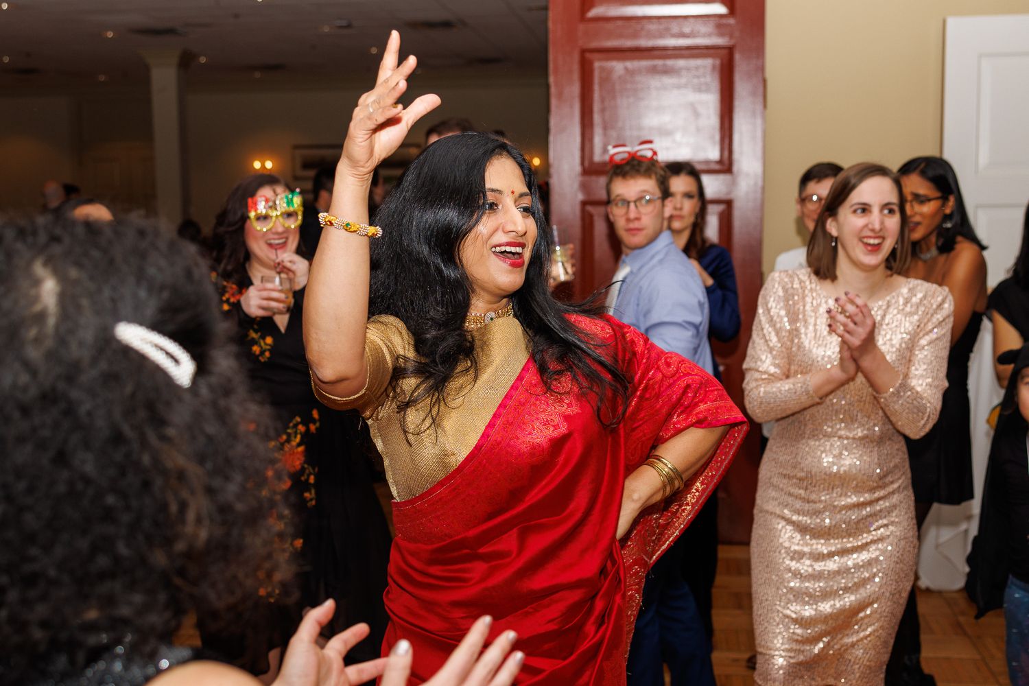 People dancing and celebrating at a wedding reception in formal attire including red and gold dresses.