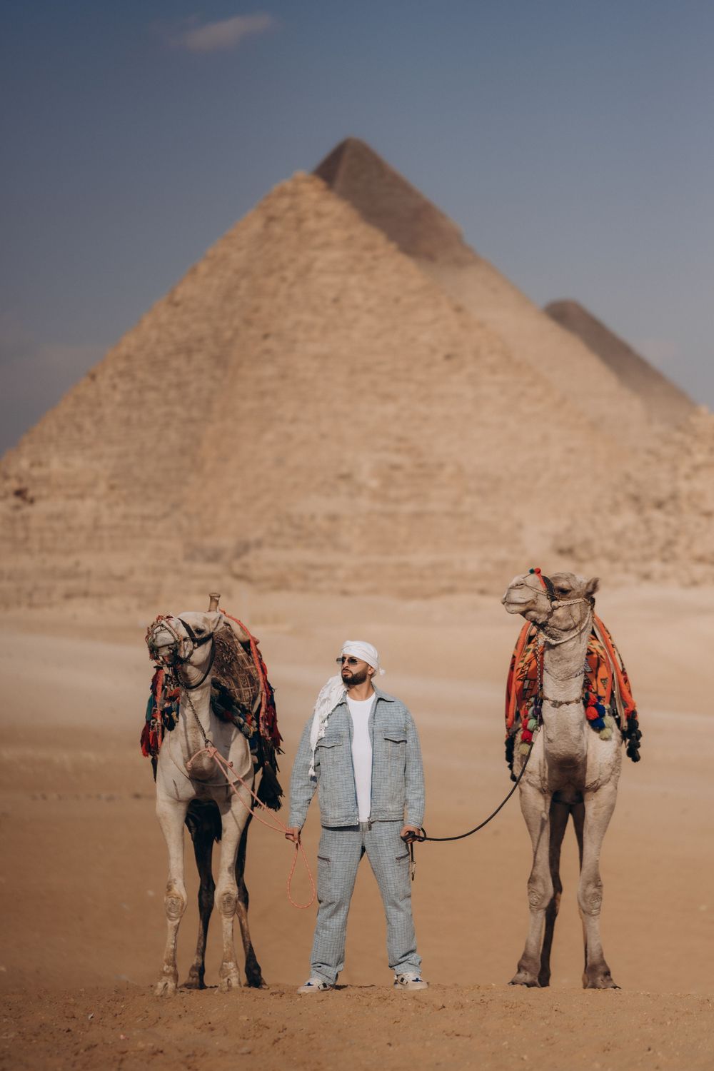 A person in light colored clothing stands between two decorated camels in front of the Great Pyramids of Giza in Egypt.