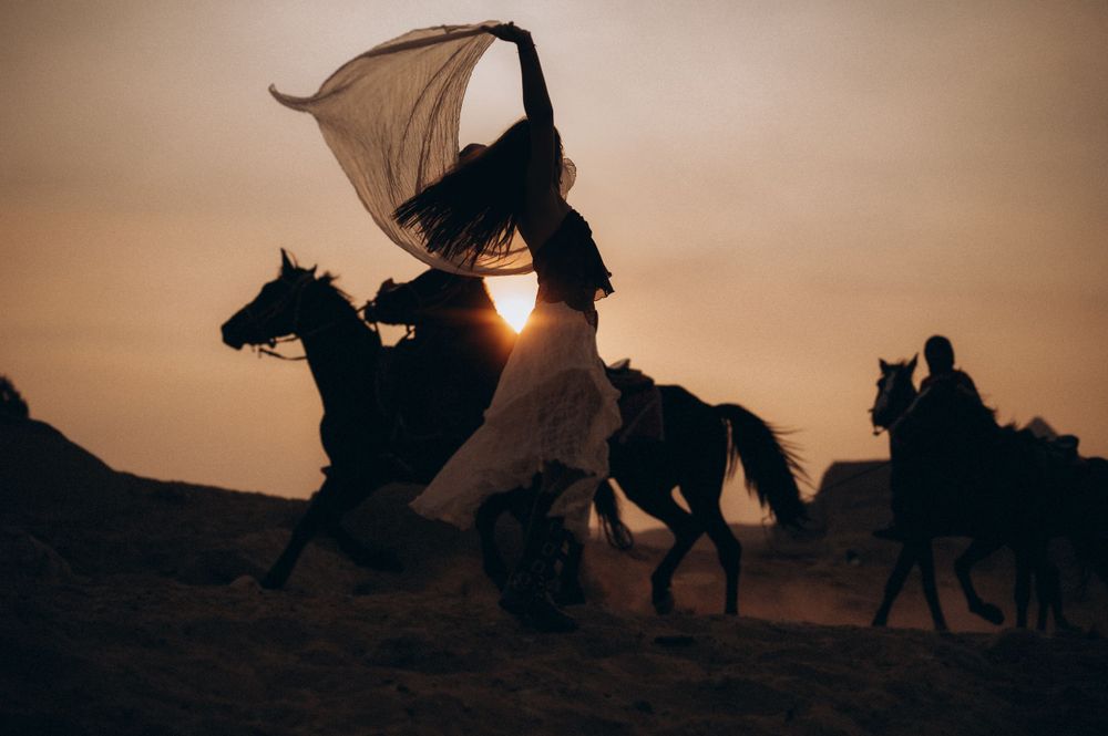 Silhouettes of horseback riders against sunset sky with flowing fabric banner creating dramatic desert scene.