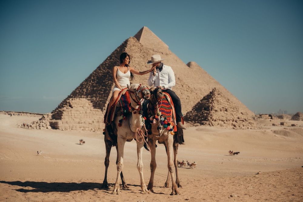 Tourists ride camels past the ancient Egyptian pyramids of Giza in the desert during golden hour.