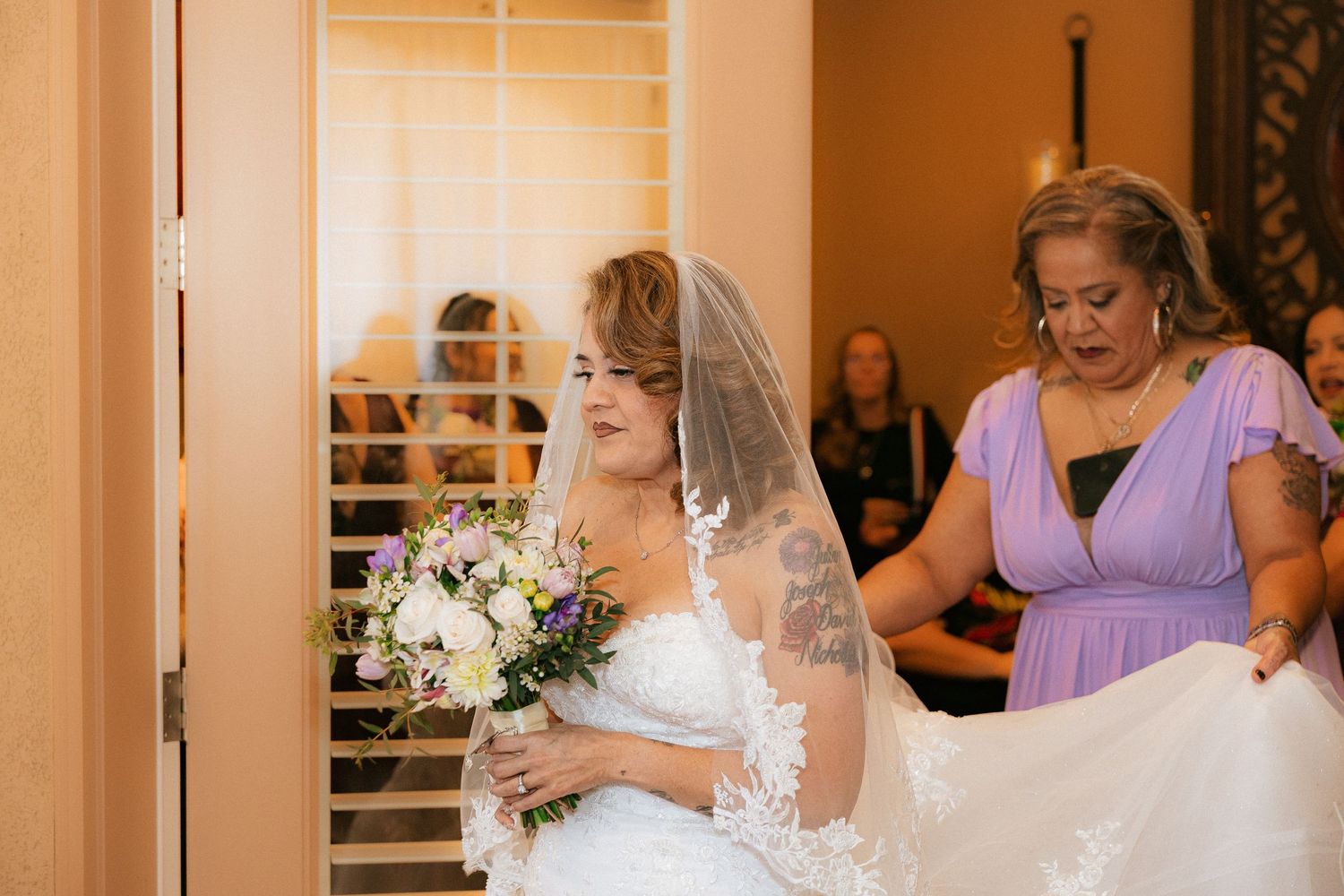 A bride in a white dress holds a bouquet while a bridesmaid in purple assists with final preparations.