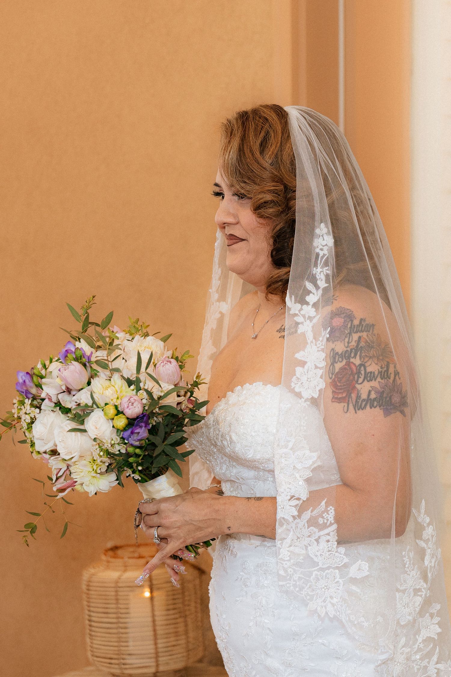 A bride in a white lace dress holds a beautiful bouquet of white and purple flowers against a peach-colored wall.