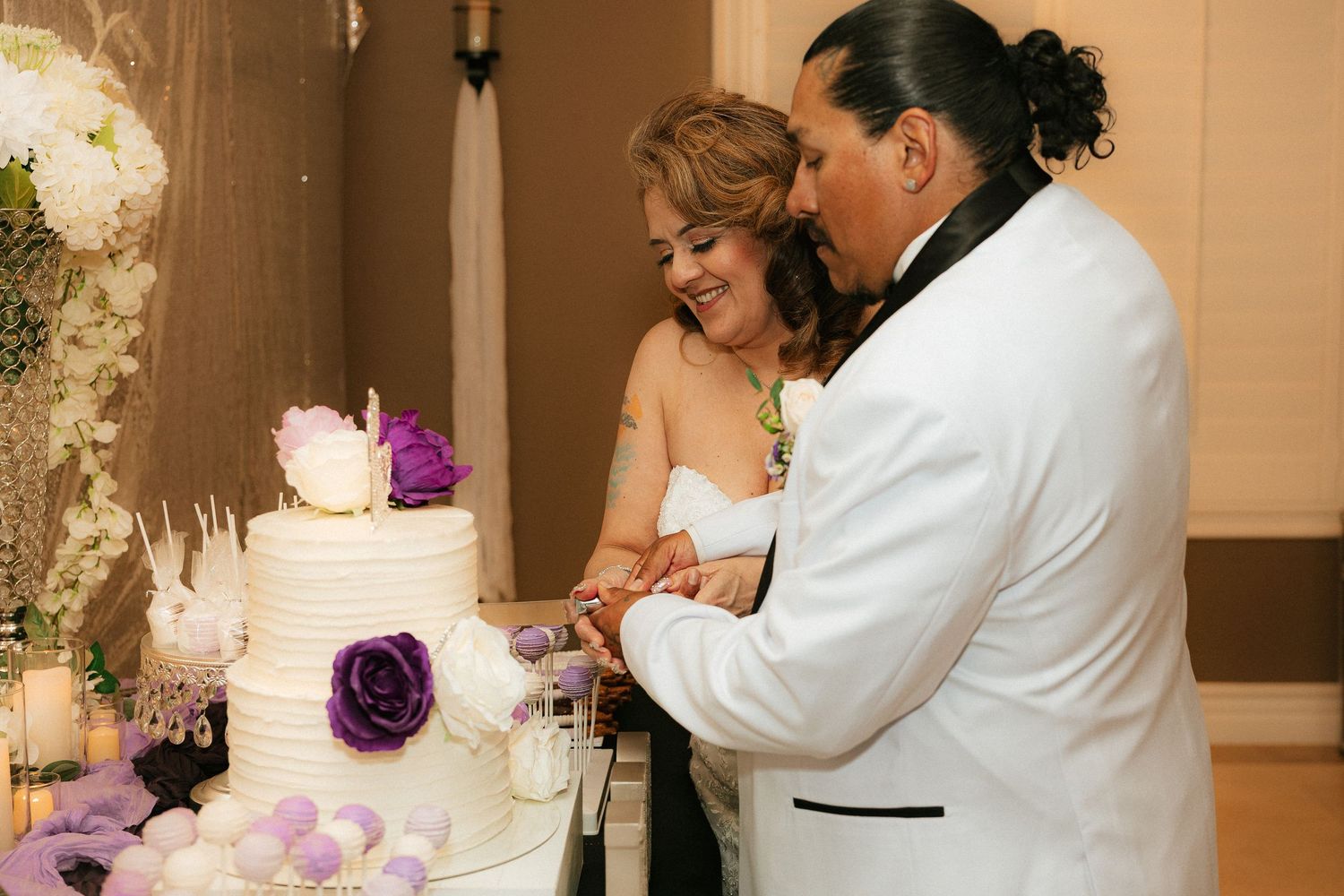 A couple wearing white attire cuts into their elegant wedding cake adorned with purple flowers at their reception.
