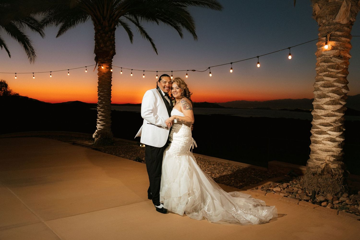 Palm trees and string lights illuminate a romantic outdoor desert wedding reception at sunset.
