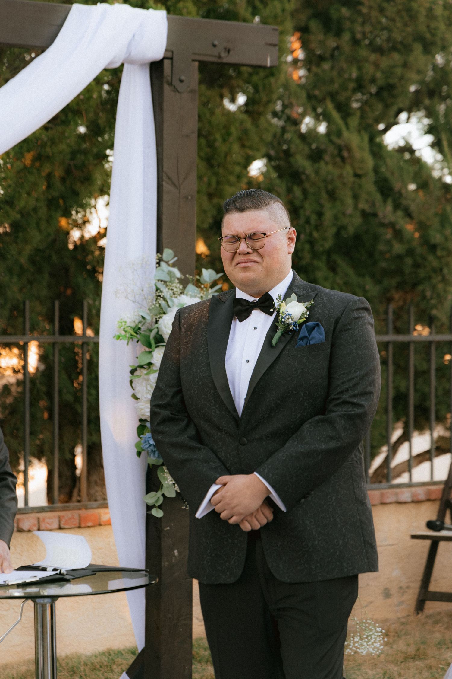 Outdoor wedding ceremony setup with wooden arch draped in white fabric against evergreen trees at sunset.