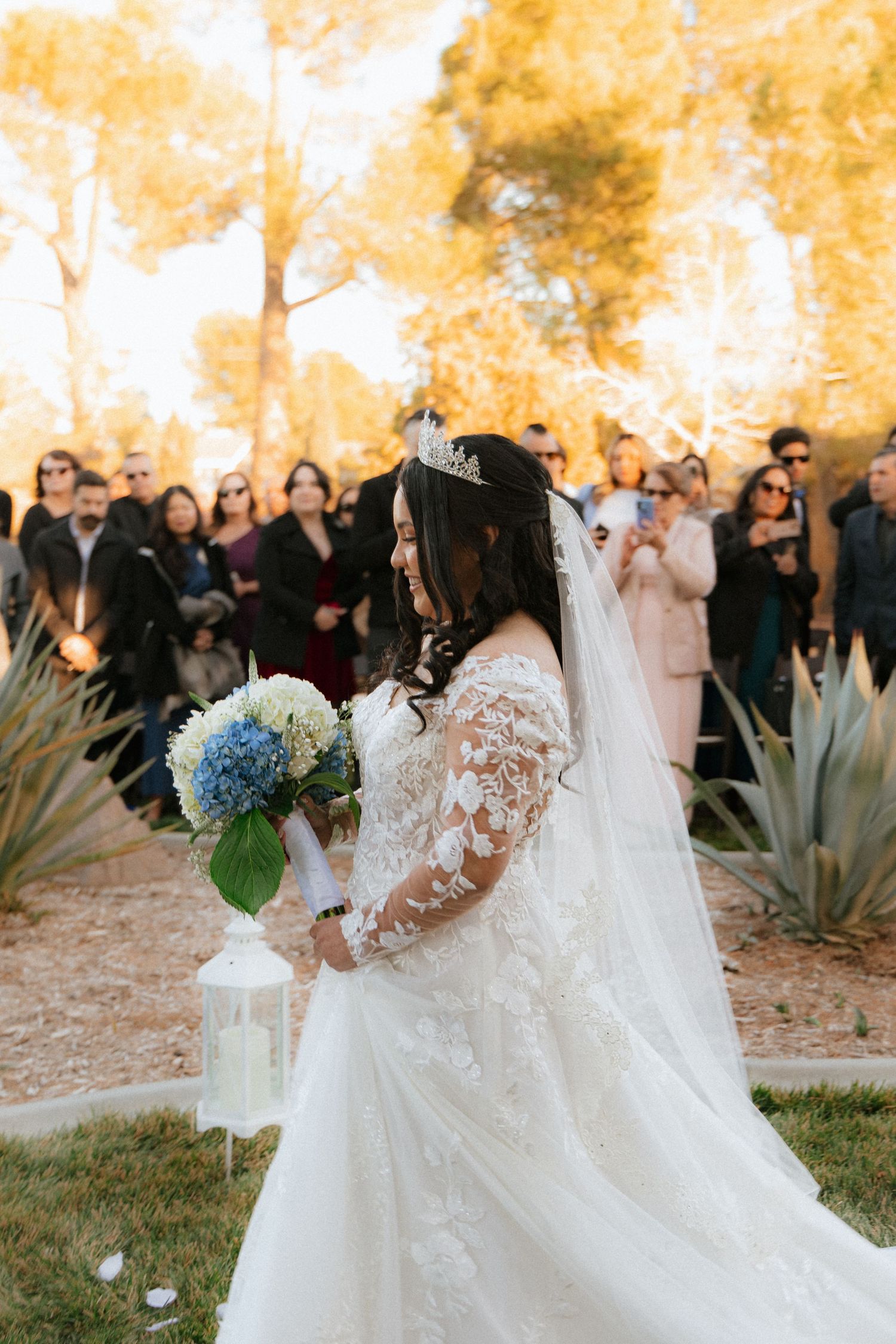 A bride in a lace long-sleeve wedding gown holds a blue hydrangea bouquet while guests look on during an autumn ceremony.