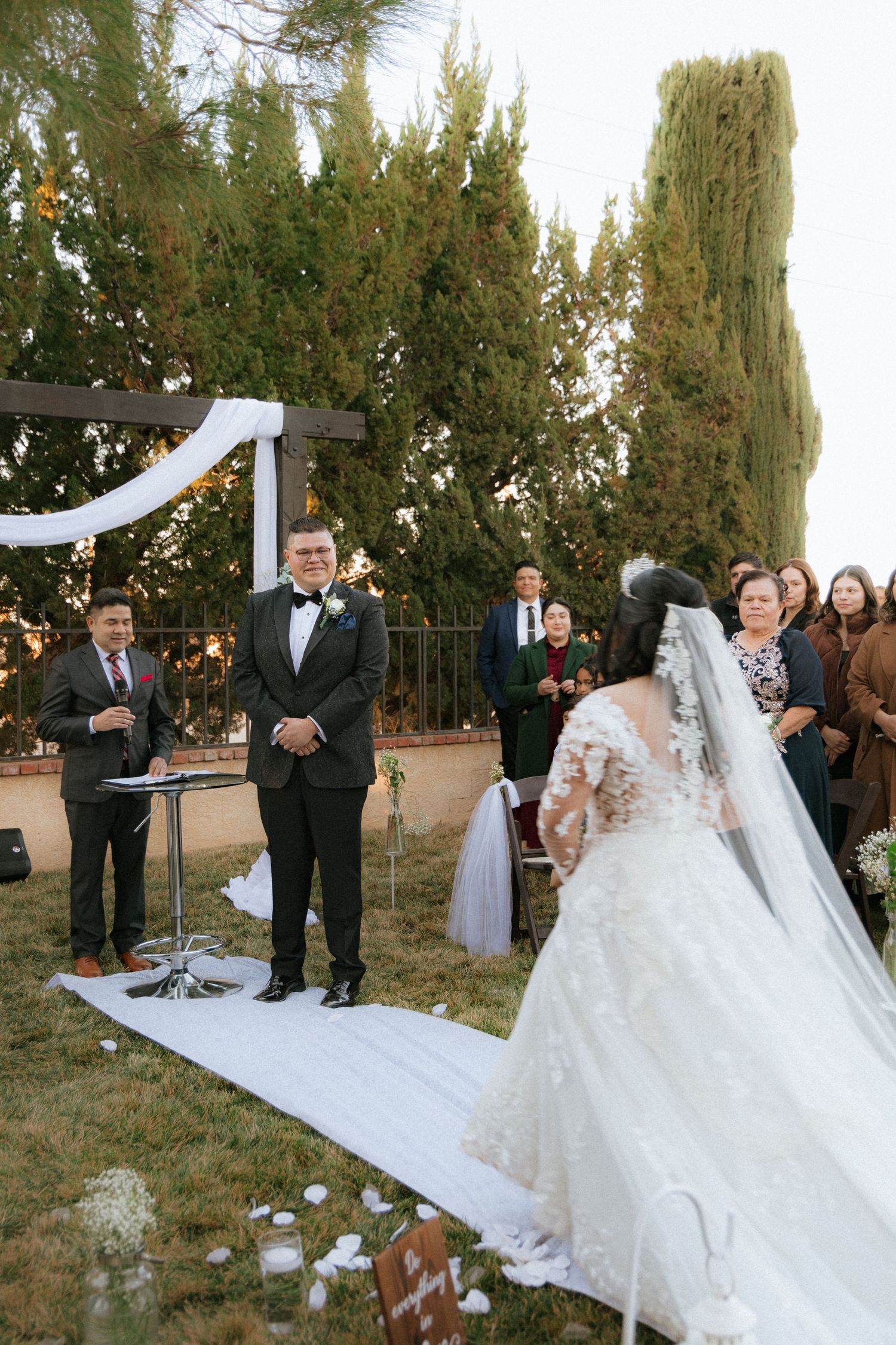 Wedding ceremony in progress under a white-draped arch with cypress trees in the background.