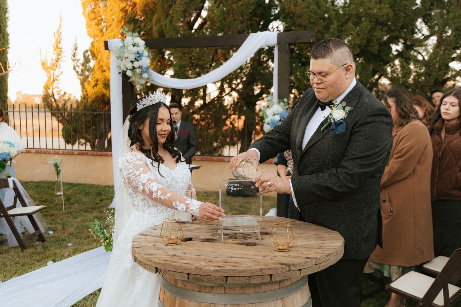 Couple performs a unity sand ceremony at outdoor wedding reception with wooden table and white decorations.