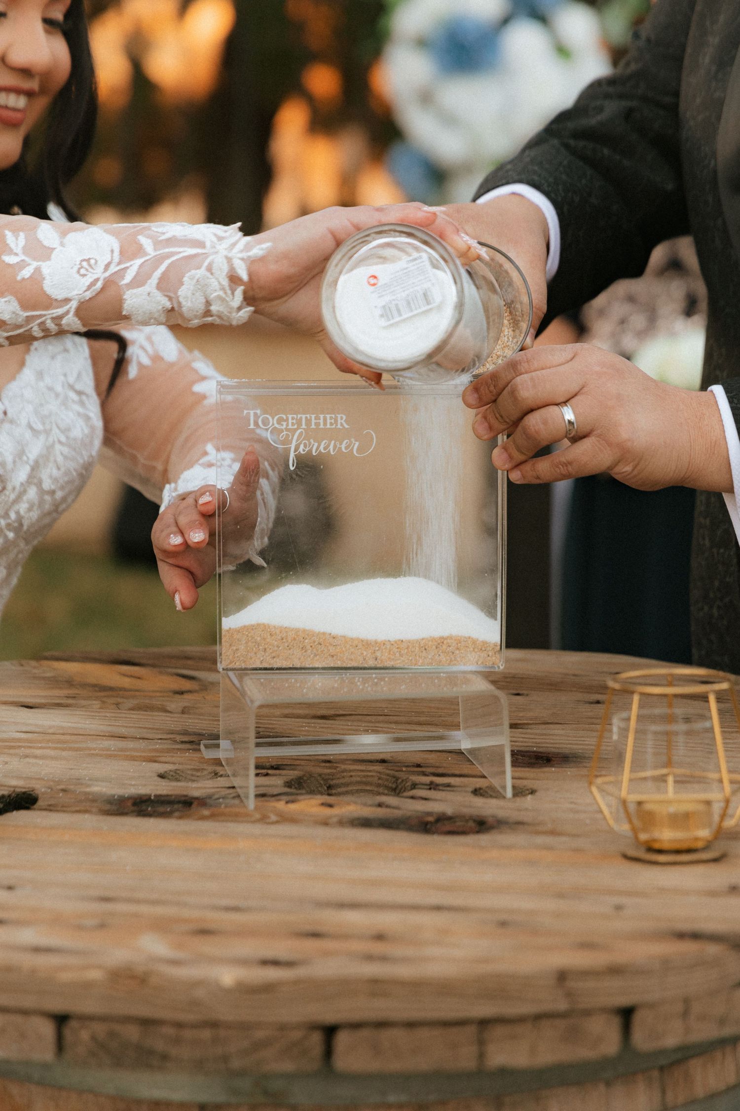 Couple pouring sand into a clear display container during a wedding unity ceremony on rustic wooden table.