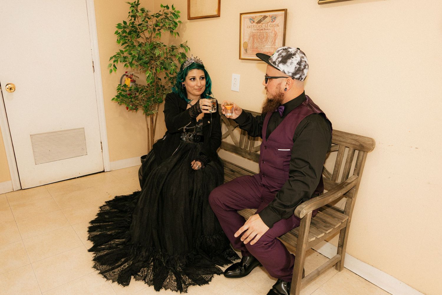 Two people sit on a wooden bench in a hallway with light beige walls and green plants in the background.