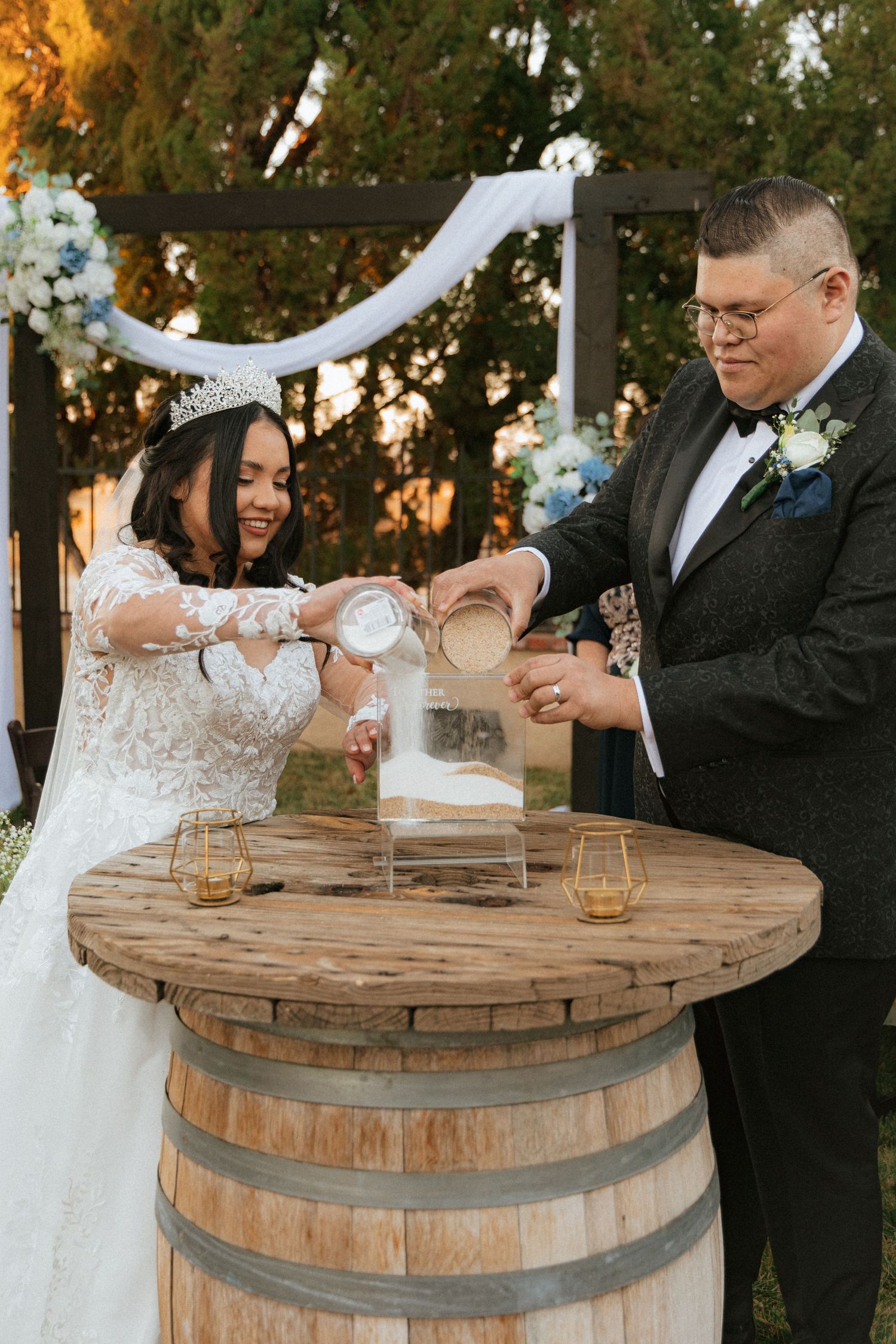 Couple participates in unity ceremony pouring ingredients into glass jar on rustic wooden barrel at sunset wedding.