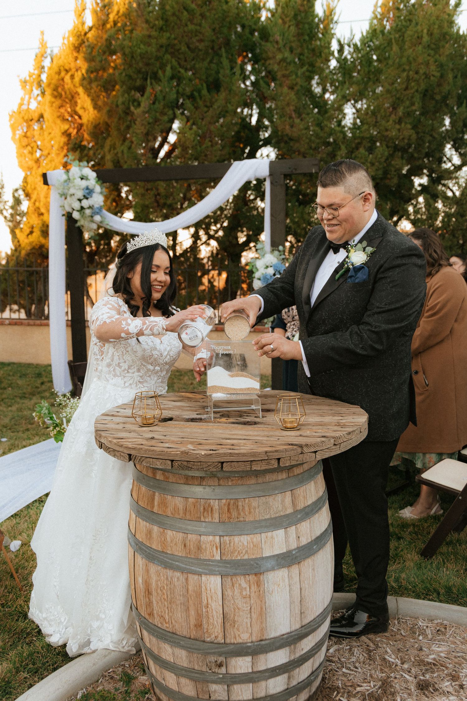Newlyweds participate in a sand ceremony at their outdoor wedding using a wooden barrel as a table.
