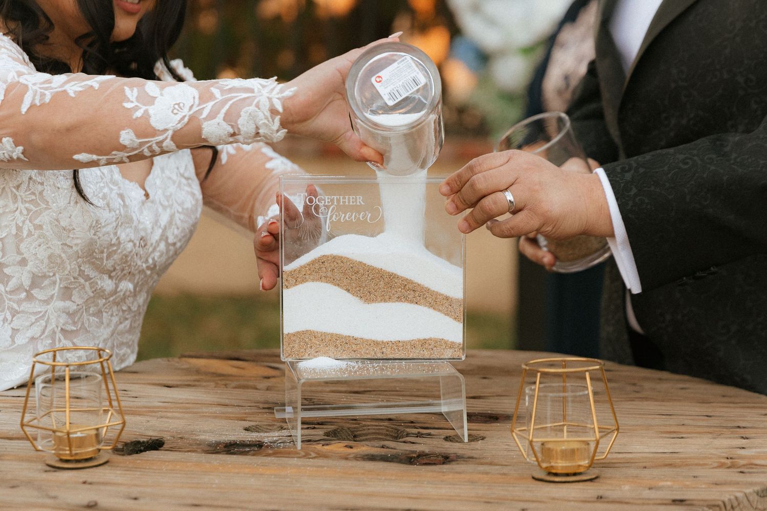 A couple performs a unity sand ceremony with glass containers and golden lanterns on a wooden table.