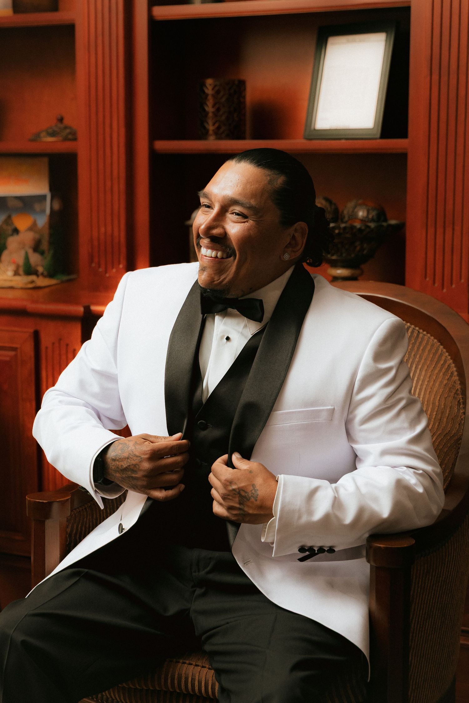A person wearing a white tuxedo jacket and black bow tie laughs joyfully while sitting in a chair near wooden bookshelves.