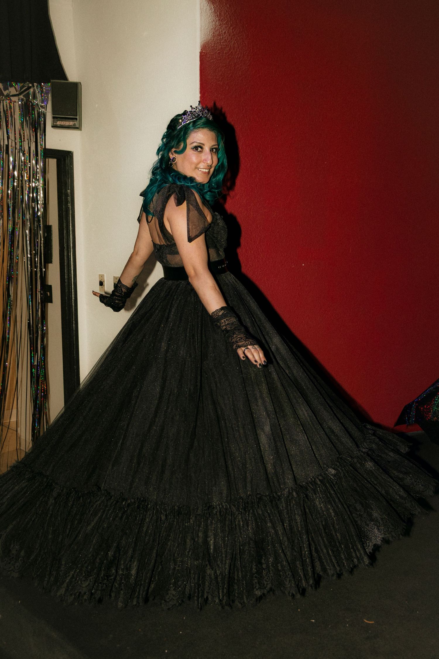 A dramatic fashion photo showcasing a flowing black gothic ball gown with long mesh sleeves against a red and white wall.