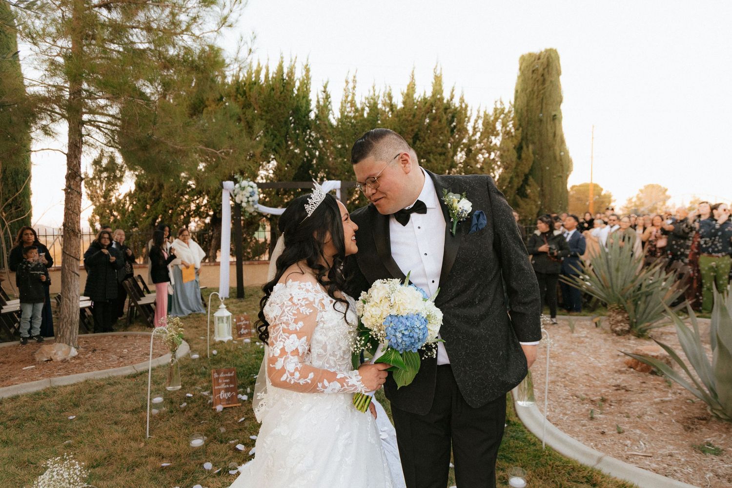 People exchange a sweet moment during a golden hour outdoor wedding ceremony surrounded by guests and trees.