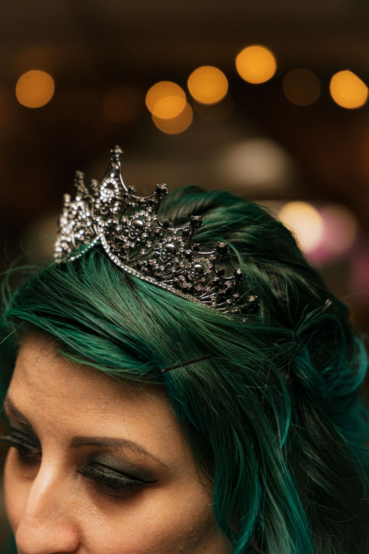 A detailed view of an ornate silver crown resting on vibrant emerald green dyed hair with warm bokeh lights in background.