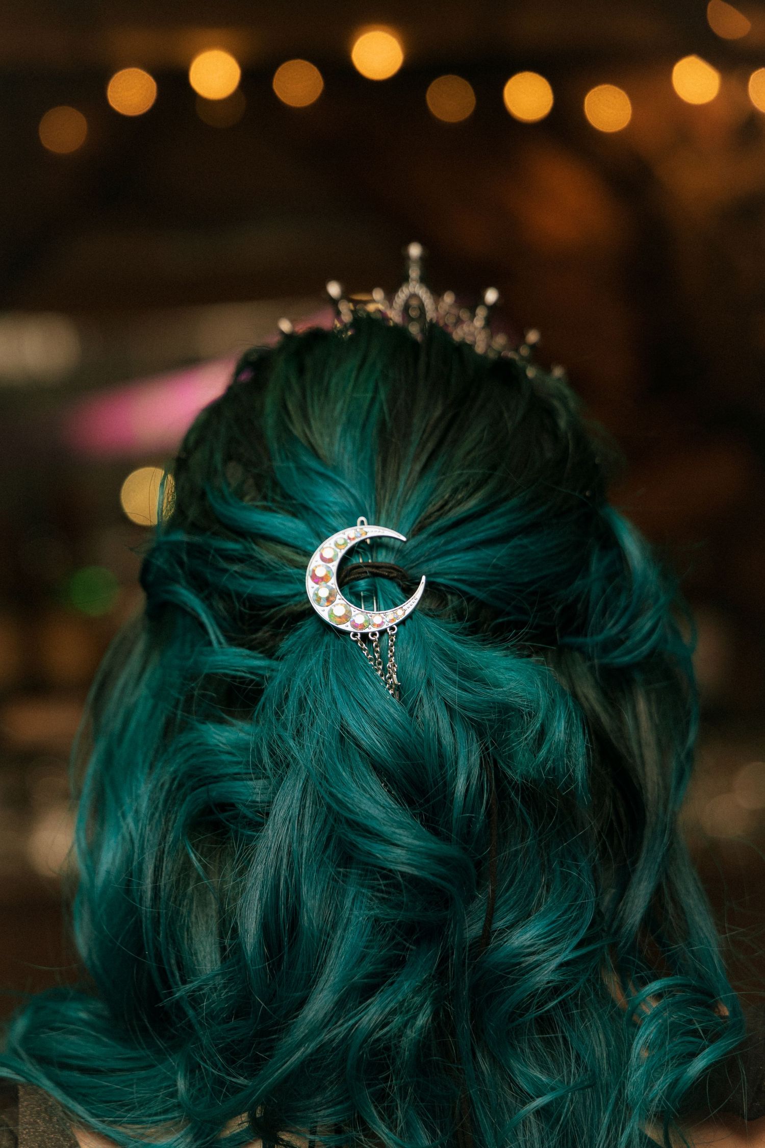 A silver crescent moon hair accessory adorns teal-dyed hair with sparkling bokeh lights creating a magical atmosphere.