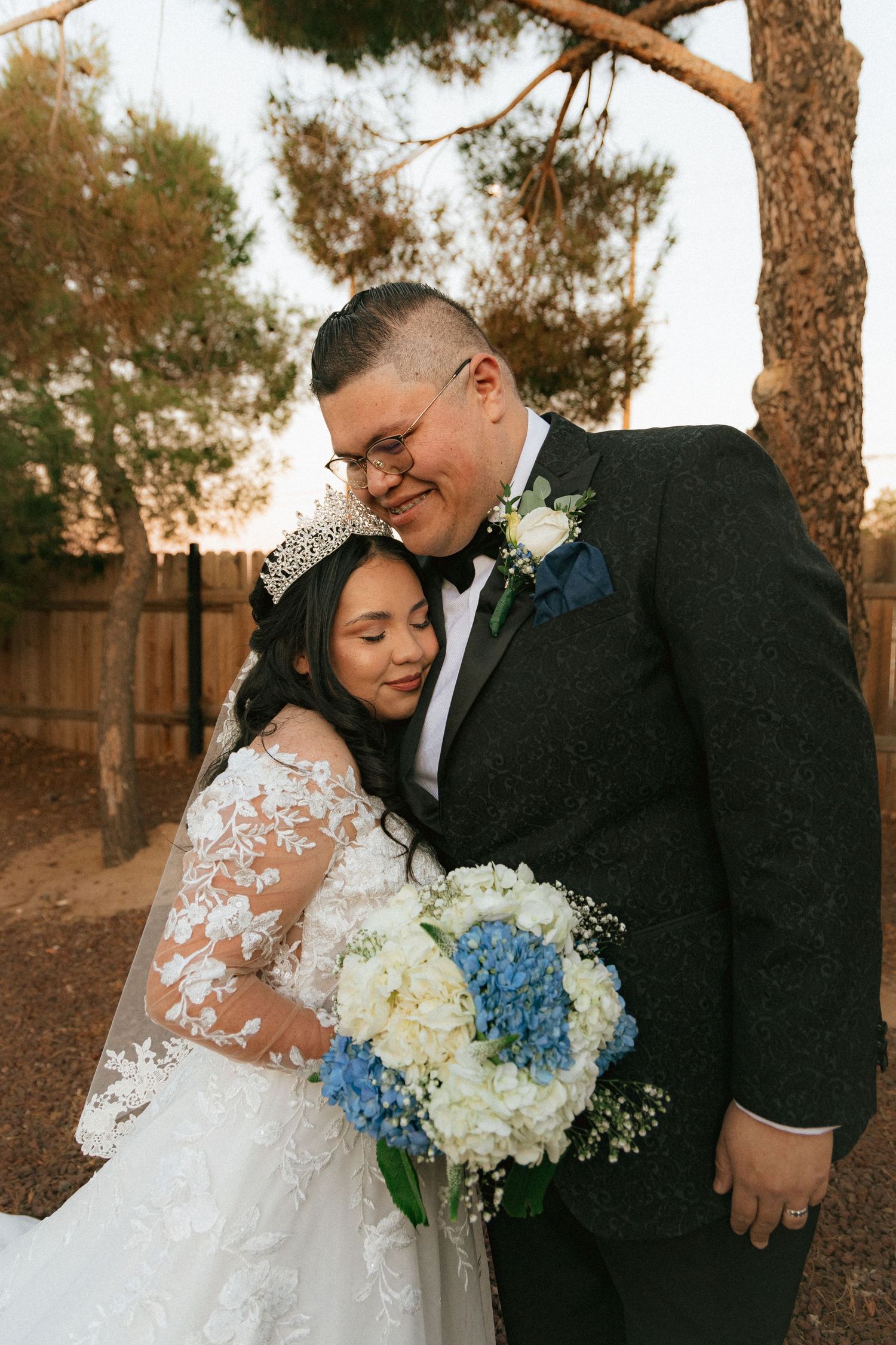 Newlyweds share intimate moment while holding blue and white floral bouquet in outdoor garden setting.