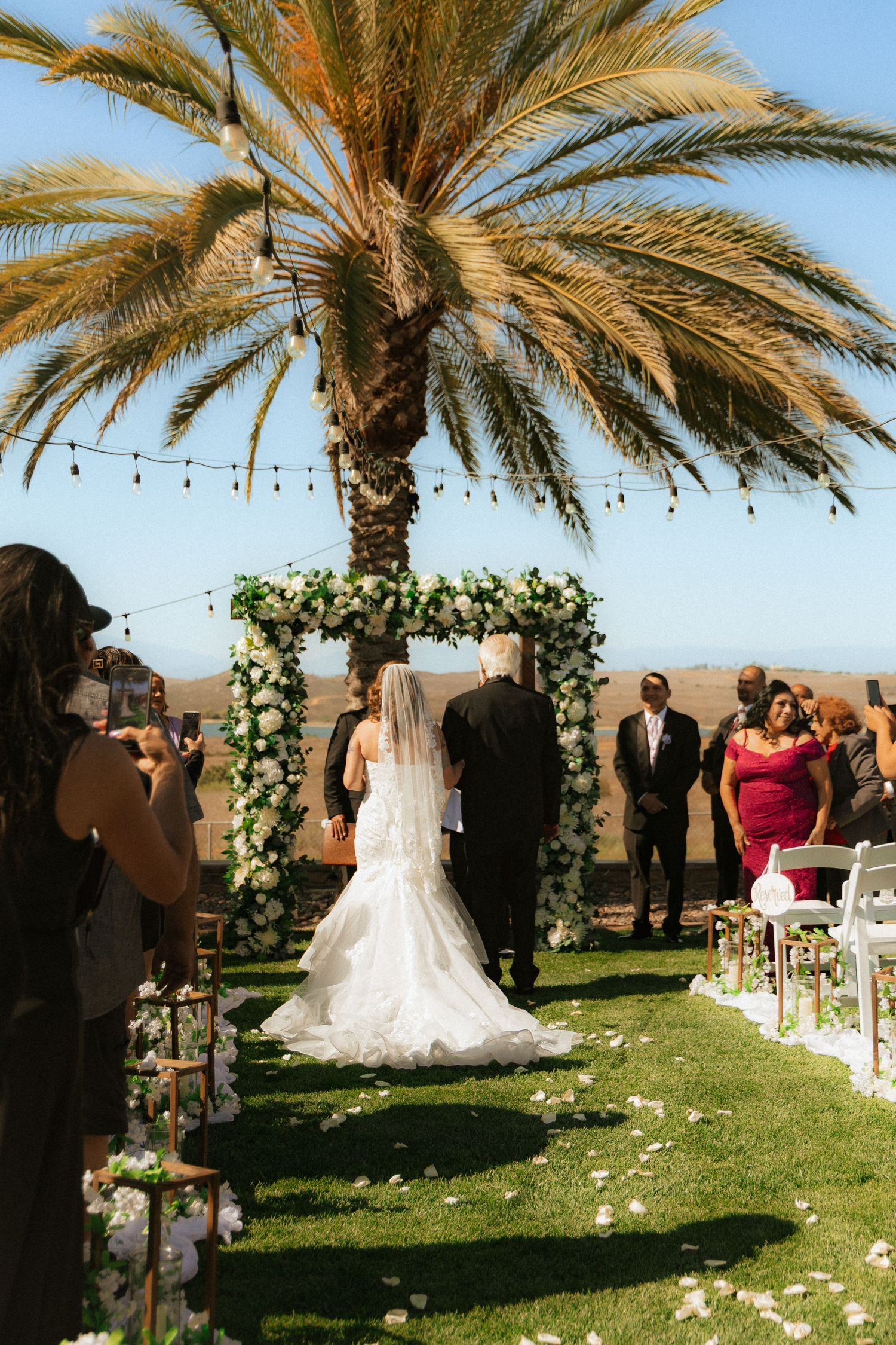 An outdoor wedding ceremony setup with palm trees, floral arch, and white petals scattered along the aisle.