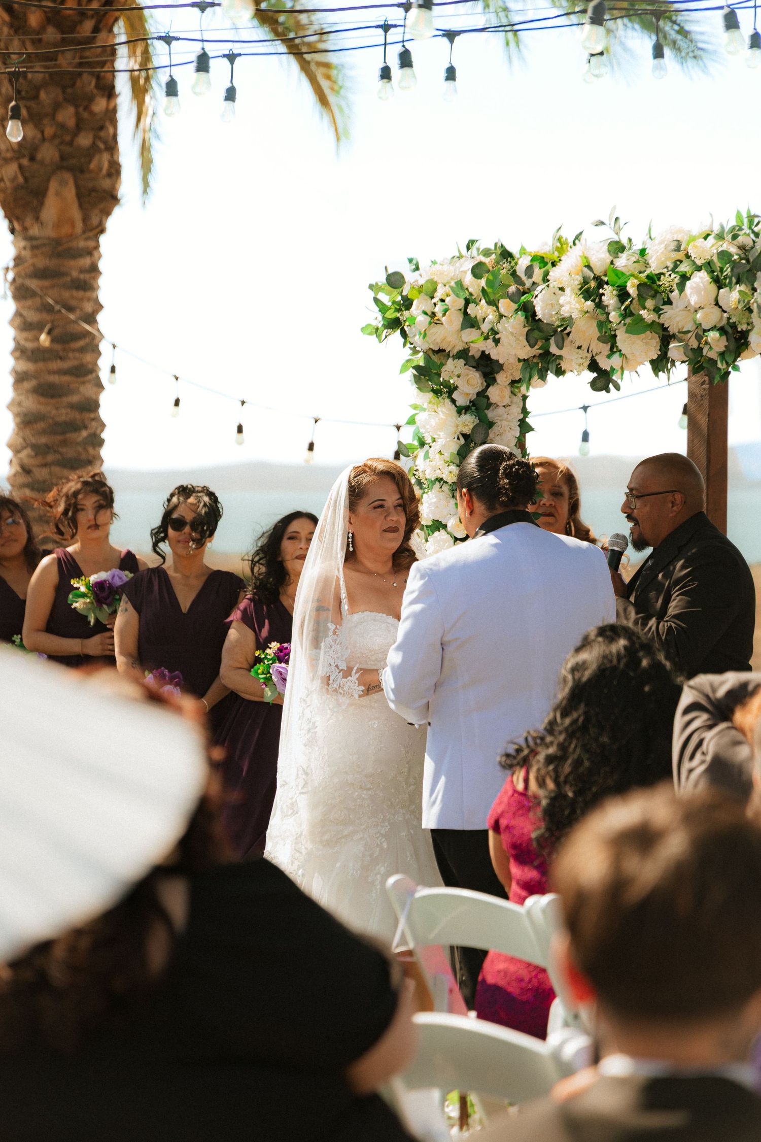 Outdoor wedding ceremony at a tropical venue with palm trees and string lights under a floral arch with white roses and greenery.