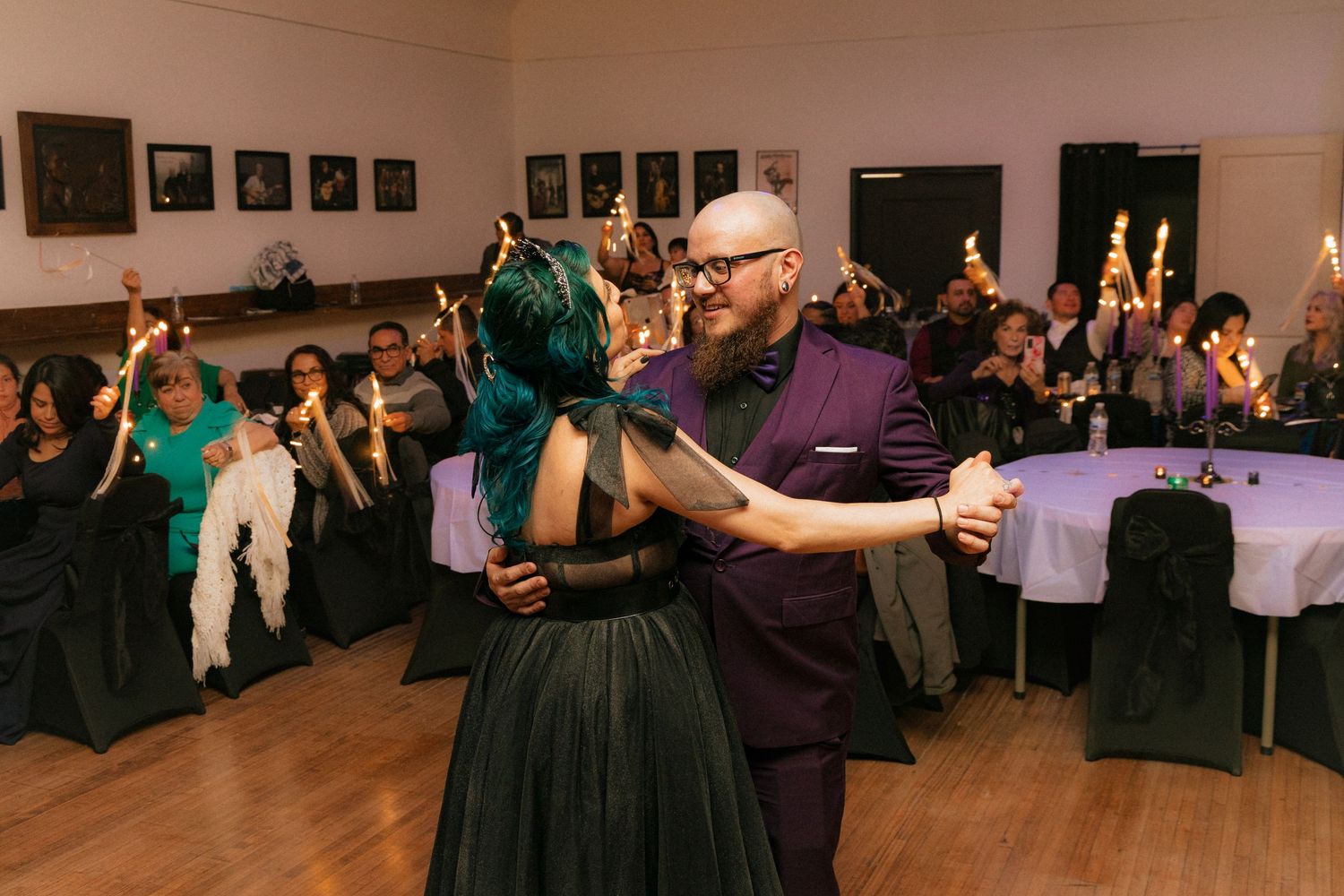 A couple dances at an indoor event venue with guests watching from decorated tables in the background.