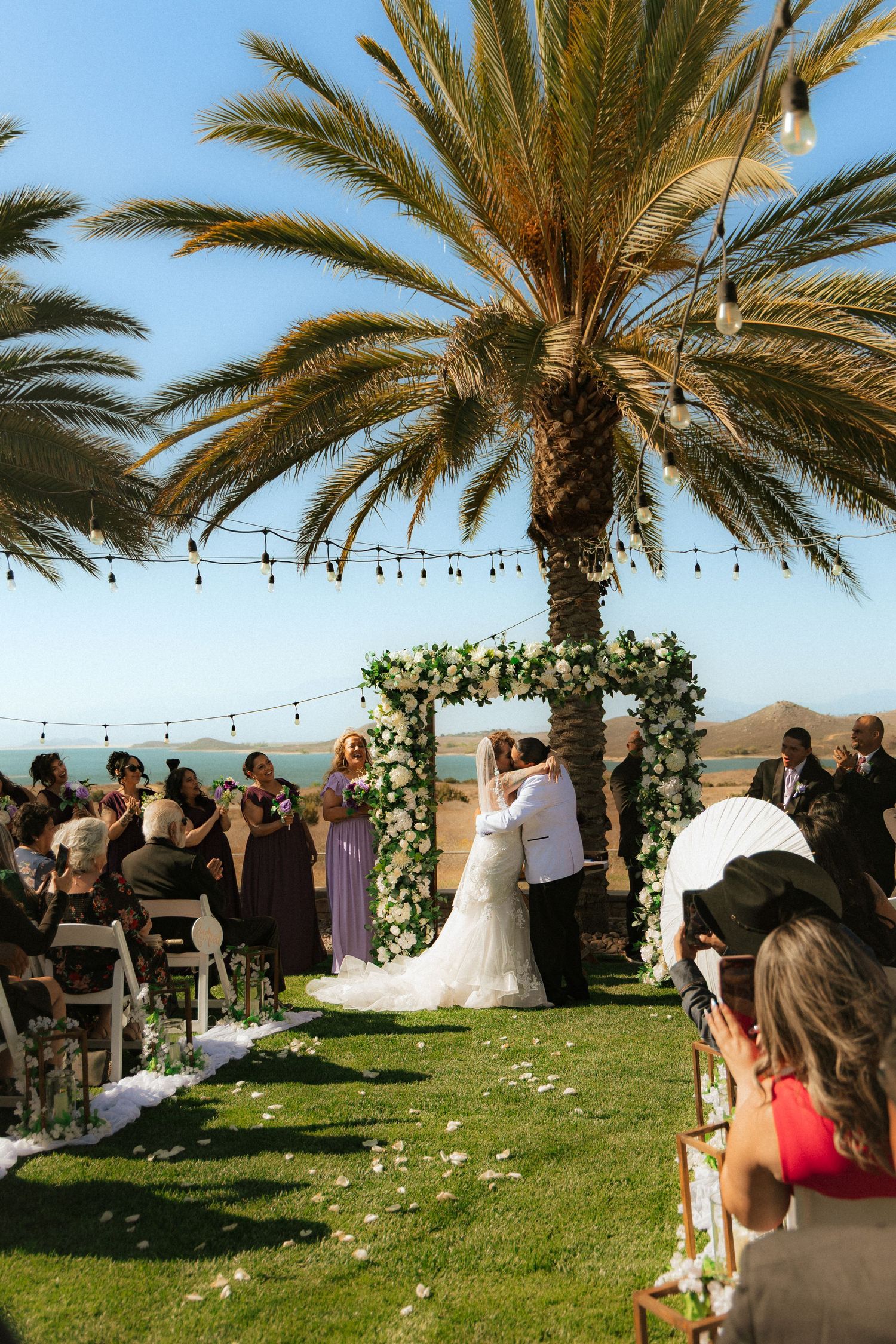 An outdoor desert wedding ceremony takes place under tall palm trees with string lights and scenic mountain views.