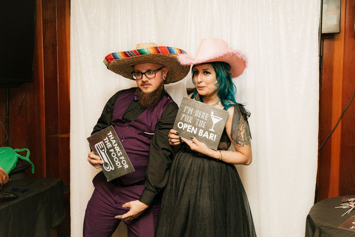 Two people in vintage-style party outfits pose together with props against white backdrop at photo booth.