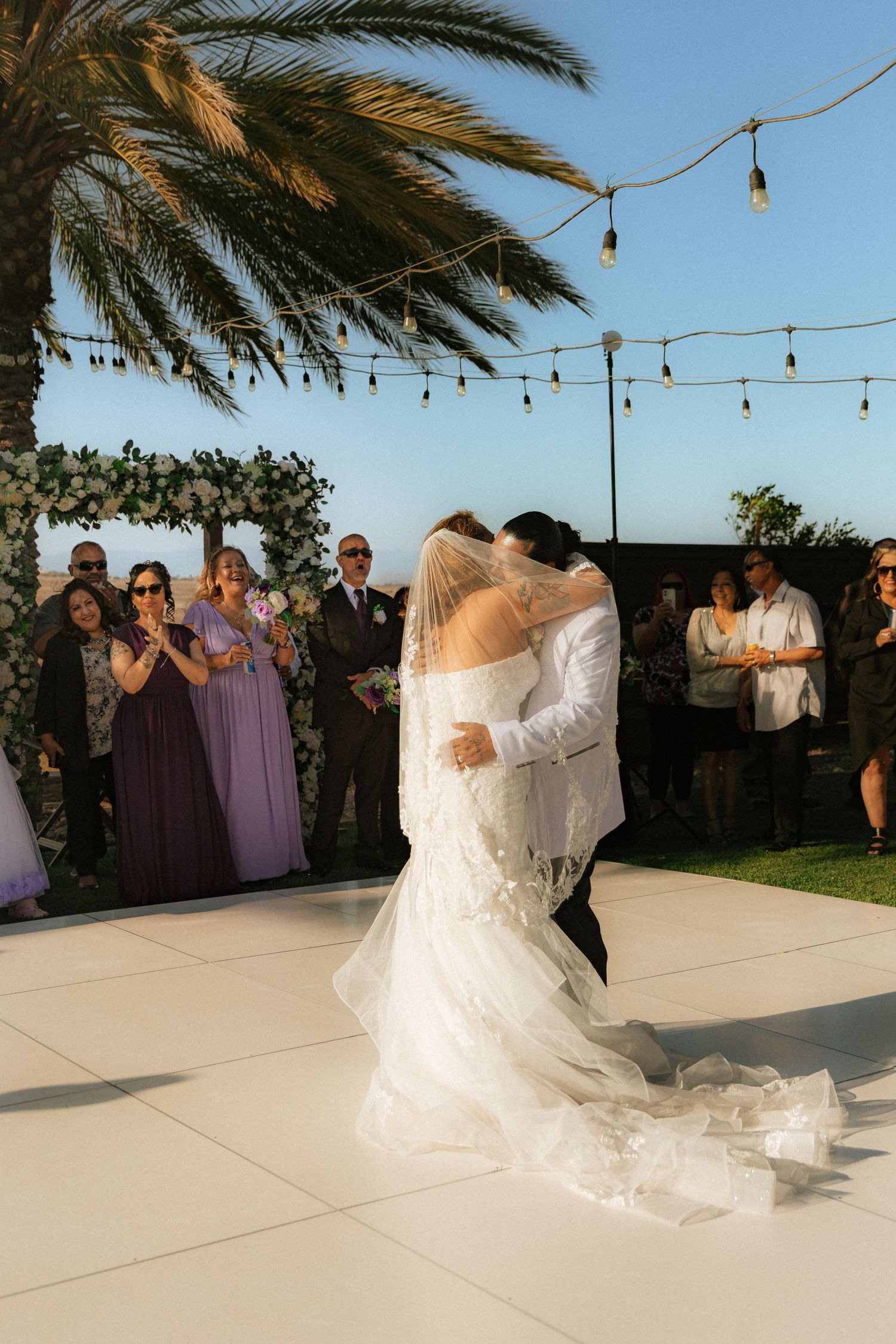 Couple shares romantic first dance at sunset outdoor wedding under string lights and palm trees.
