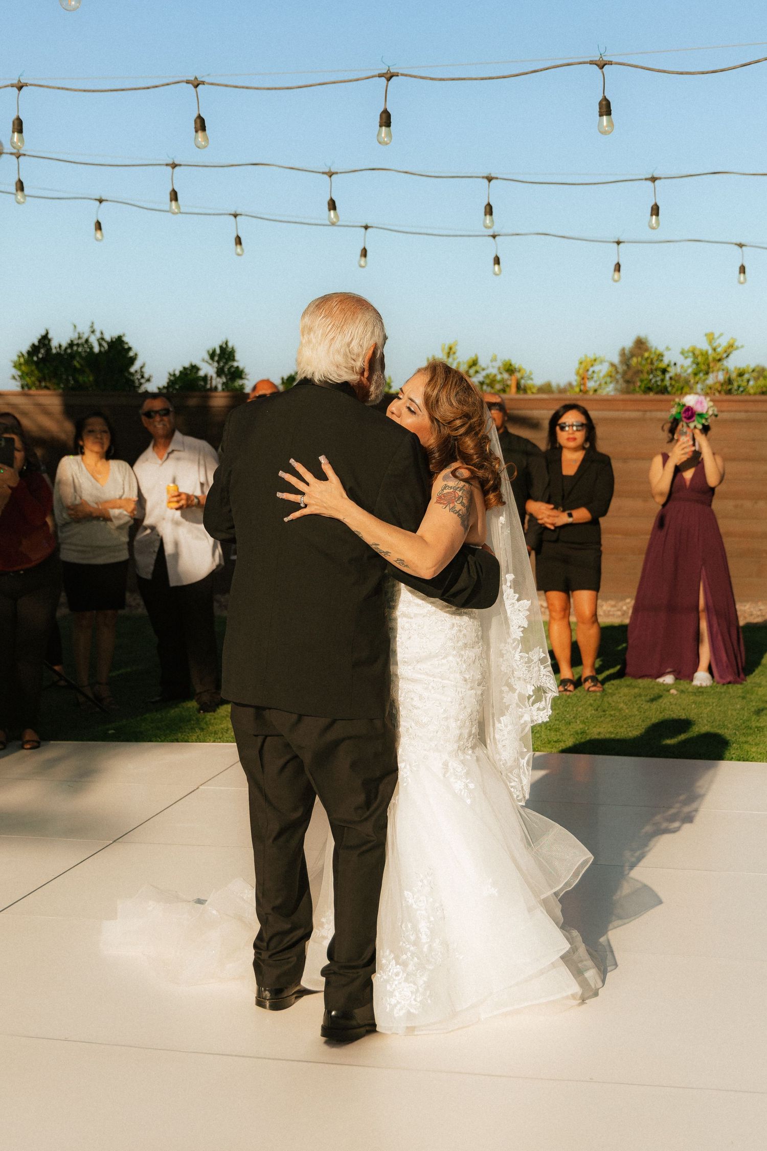 A couple dances together outdoors at sunset under strung lights while guests watch at a wedding reception.
