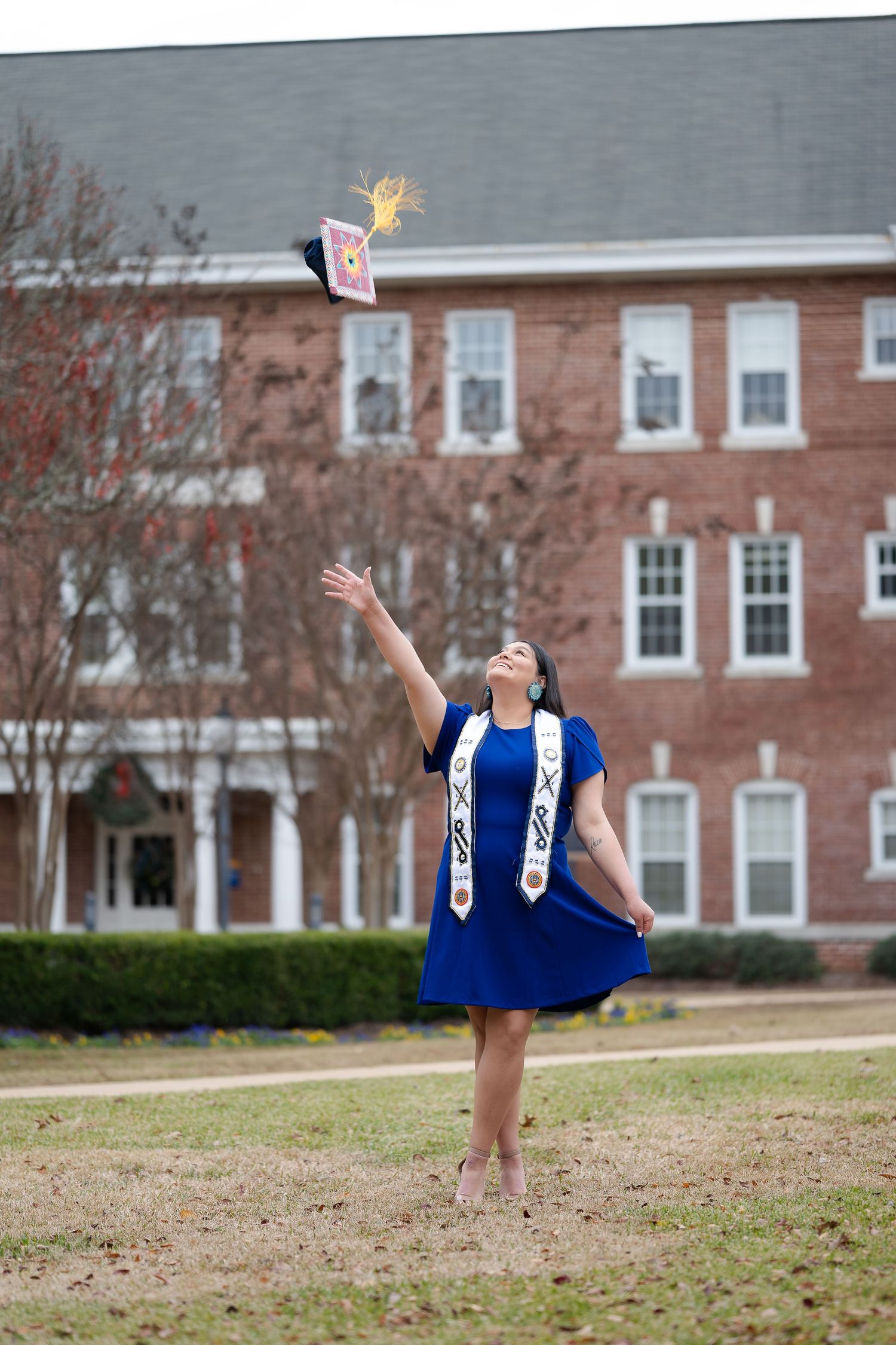 Graduate in blue dress and ceremonial stole tosses cap into air on college campus.